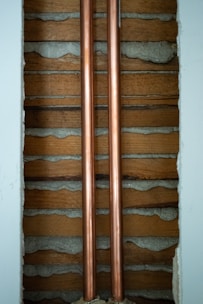 Two copper pipes run vertically between wooden slats and concrete. The pipes are shiny and metallic, while the surrounding wall has a rustic look with exposed wood and cement.