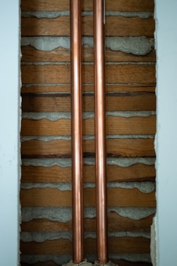 Two copper pipes run vertically between wooden slats and concrete. The pipes are shiny and metallic, while the surrounding wall has a rustic look with exposed wood and cement.