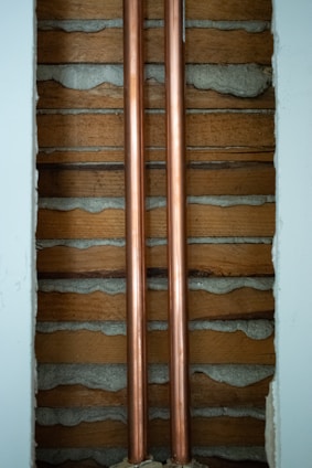 Two copper pipes run vertically between wooden slats and concrete. The pipes are shiny and metallic, while the surrounding wall has a rustic look with exposed wood and cement.