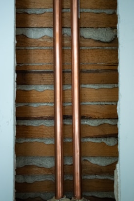 Two copper pipes run vertically between wooden slats and concrete. The pipes are shiny and metallic, while the surrounding wall has a rustic look with exposed wood and cement.