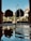 A serene courtyard features a central fountain with water gently streaming into a reflective pool. The scene is framed by lush greenery, tall trimmed shrubs, and an impressive architectural backdrop with arches. Sunlight casts soft shadows, creating a peaceful and harmonious atmosphere.