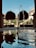 A serene courtyard features a central fountain with water gently streaming into a reflective pool. The scene is framed by lush greenery, tall trimmed shrubs, and an impressive architectural backdrop with arches. Sunlight casts soft shadows, creating a peaceful and harmonious atmosphere.