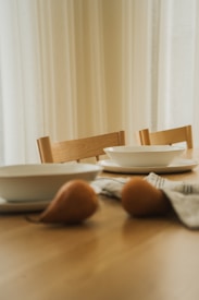 A calm dining scene featuring a wooden table set with simple white plates and bowls. Wooden chairs are neatly placed around the table, and two pears lie on the table alongside a folded cloth napkin. The setting is softly illuminated by natural light filtering through sheer curtains.