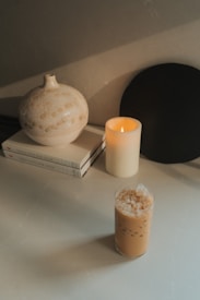 A serene tabletop setting features a round ceramic vase with a speckled pattern, two stacked books with a minimalist design, an ivory-colored candle softly lit, and a glass of iced coffee filled with ice cubes. The overall aesthetic is calming and composed.