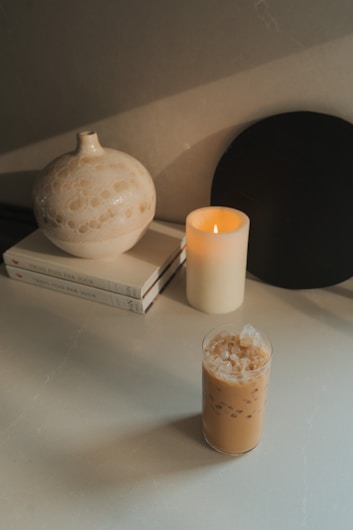 A serene tabletop setting features a round ceramic vase with a speckled pattern, two stacked books with a minimalist design, an ivory-colored candle softly lit, and a glass of iced coffee filled with ice cubes. The overall aesthetic is calming and composed.