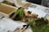 A collection of terrarium-making materials is arranged on a table, including a tray filled with soil, pieces of moss, various sizes of stones, and gardening tools. Sheets with printed text and images are partially obscured by the items, suggesting instructions or guidelines.