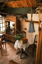 A cozy rustic kitchen scene featuring a small round table covered with a checkered cloth, on which a wooden crate with pumpkins and a vase of flowers are placed. Surrounding the table are wooden chairs. In the background, there is an antique wooden cabinet with glass doors displaying dishes inside. The room has a brick floor and a wooden ceiling, with a hanging metal bell in the foreground. There is natural light streaming in from a large window to the left.