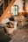 A cozy living space featuring wooden stairs and floors, with a rustic theme. Gingham-patterned couches sit against a wooden wall, with a green-framed window between them. A basket filled with pinecones and miniature pumpkins is placed near the stairs, enhancing the autumnal and homey feel of the room.
