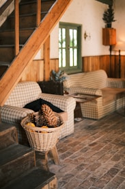 A cozy living space featuring wooden stairs and floors, with a rustic theme. Gingham-patterned couches sit against a wooden wall, with a green-framed window between them. A basket filled with pinecones and miniature pumpkins is placed near the stairs, enhancing the autumnal and homey feel of the room.