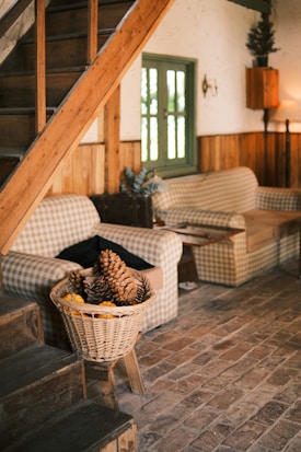 A cozy living space featuring wooden stairs and floors, with a rustic theme. Gingham-patterned couches sit against a wooden wall, with a green-framed window between them. A basket filled with pinecones and miniature pumpkins is placed near the stairs, enhancing the autumnal and homey feel of the room.