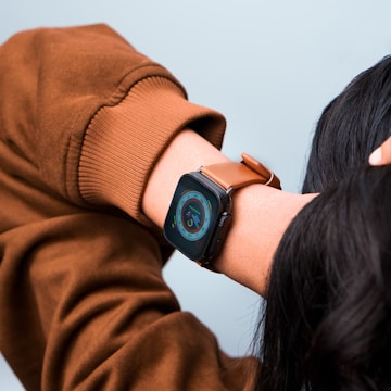 A person wearing a smartwatch with a brown leather strap. The smartwatch displays a colorful interface with various icons and a circular dial. The person is dressed in a brown long-sleeve top, and their hand is raised over their head.