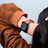 A person wearing a smartwatch with a brown leather strap. The smartwatch displays a colorful interface with various icons and a circular dial. The person is dressed in a brown long-sleeve top, and their hand is raised over their head.
