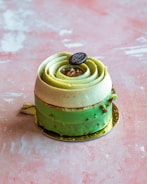 A decorative pastry shaped like a rose, featuring layers of light green and cream colors. It's topped with a small round button-like decoration, placed on a light pink textured surface.