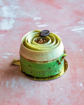 A decorative pastry shaped like a rose, featuring layers of light green and cream colors. It's topped with a small round button-like decoration, placed on a light pink textured surface.