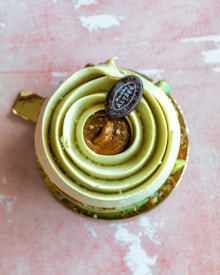 A beautifully crafted dessert featuring layered spirals of green cream surrounding a caramel-colored center, topped with a chocolate disc bearing the label 'MAISON ALZEE'. The dessert sits on a textured pink surface, and its base is garnished with small bits of what seems to be nuts or similar toppings.