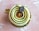 A beautifully crafted pastry with spiral layers of green cream and a glossy caramel center. Topped with a small oval chocolate plaque featuring delicate text. The dessert is placed on a golden base, set against a textured pink background.