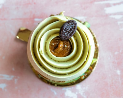 A beautifully crafted pastry with spiral layers of green cream and a glossy caramel center. Topped with a small oval chocolate plaque featuring delicate text. The dessert is placed on a golden base, set against a textured pink background.