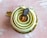 A beautifully crafted pastry with spiral layers of green cream and a glossy caramel center. Topped with a small oval chocolate plaque featuring delicate text. The dessert is placed on a golden base, set against a textured pink background.
