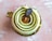 A beautifully crafted pastry with spiral layers of green cream and a glossy caramel center. Topped with a small oval chocolate plaque featuring delicate text. The dessert is placed on a golden base, set against a textured pink background.
