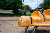 A bright yellow playground seesaw shaped like a bee is situated on a sandy surface. It features black handlebars and large cartoonish eyes. A wooden bench is visible in the background, surrounded by green trees and an overcast sky.