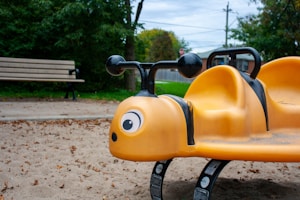 A bright yellow playground seesaw shaped like a bee is situated on a sandy surface. It features black handlebars and large cartoonish eyes. A wooden bench is visible in the background, surrounded by green trees and an overcast sky.