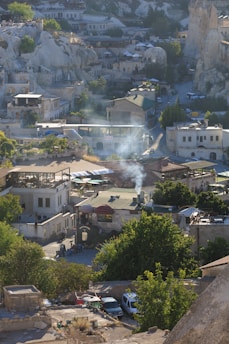 A picturesque town with buildings nestled among unique rock formations. Smoke rises from a chimney, adding character to the rustic architecture. Lush greenery contrasts with the stone structures, and cars are parked along narrow streets, creating a serene village atmosphere.