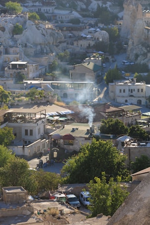 A picturesque town with buildings nestled among unique rock formations. Smoke rises from a chimney, adding character to the rustic architecture. Lush greenery contrasts with the stone structures, and cars are parked along narrow streets, creating a serene village atmosphere.