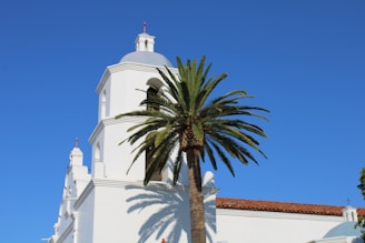 A white mission-style building with a bell tower is positioned next to a tall palm tree under a clear blue sky. The structure features arches and red-tiled roofing, typical of Spanish colonial architecture.