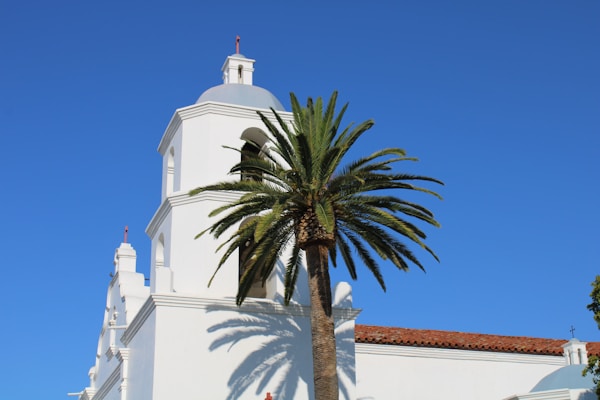 A white mission-style building with a bell tower is positioned next to a tall palm tree under a clear blue sky. The structure features arches and red-tiled roofing, typical of Spanish colonial architecture.