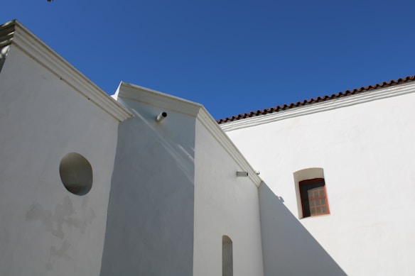 A building facade featuring white stucco walls and a terracotta-tiled roof set against a clear blue sky. Architectural elements include geometric shapes, a circular window, a rectangular window with a grid pattern, and a cylindrical protrusion.