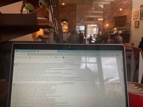 A laptop screen displaying a webpage is in the foreground, within a cozy cafe setting. The background shows a rustic interior with exposed brick walls, potted plants, and soft lighting. Patrons are seated at tables, engaged in conversation, creating a relaxed atmosphere.