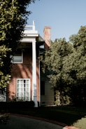 A brick house partially covered by dense greenery, with a white column supporting a porch roof. Sunlight casts shadows against the wall, and a chimney is visible above the roofline. The windows are covered by white curtains, and the lawn is well-maintained with a stone pathway leading to the entrance.