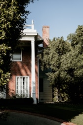 A brick house partially covered by dense greenery, with a white column supporting a porch roof. Sunlight casts shadows against the wall, and a chimney is visible above the roofline. The windows are covered by white curtains, and the lawn is well-maintained with a stone pathway leading to the entrance.