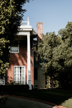 A brick house partially covered by dense greenery, with a white column supporting a porch roof. Sunlight casts shadows against the wall, and a chimney is visible above the roofline. The windows are covered by white curtains, and the lawn is well-maintained with a stone pathway leading to the entrance.