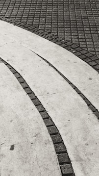 A textured pavement pattern featuring alternating curved lighter and darker surfaces, with small square tiles forming a grid in the darker areas and a smooth concrete finish on the lighter sections. The contrast creates a sense of movement and rhythm.