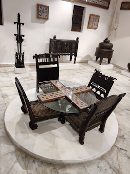 A room features a central round glass table with four intricately designed wooden chairs placed on a circular platform. The chairs have wicker seats and detailed carvings. The background includes a decorative wooden cabinet, a tall metal stand, and an antique-looking pot on stands, all set against white walls with framed artwork and small square windows.