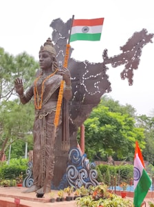 A large bronze statue of a figure adorned with intricate jewelry and garlands stands in front of a metal sculpture depicting the map of India. The figure holds the Indian national flag in one hand. Surrounding the statue are lush green trees and neatly arranged potted plants.