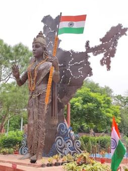 A large bronze statue of a figure adorned with intricate jewelry and garlands stands in front of a metal sculpture depicting the map of India. The figure holds the Indian national flag in one hand. Surrounding the statue are lush green trees and neatly arranged potted plants.
