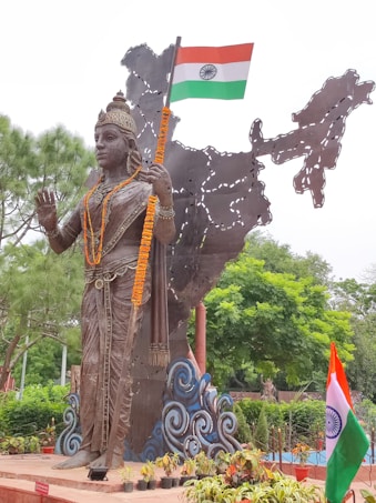 A large bronze statue of a figure adorned with intricate jewelry and garlands stands in front of a metal sculpture depicting the map of India. The figure holds the Indian national flag in one hand. Surrounding the statue are lush green trees and neatly arranged potted plants.