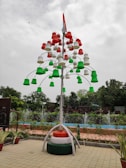 A creatively designed artificial tree made up of colorful pot-like structures in red, white, and green is displayed in an outdoor setting. It is topped with a national flag. The background features lush greenery, fountains, and sculpted panels. Potted plants are arranged at the base, while the sky appears overcast.
