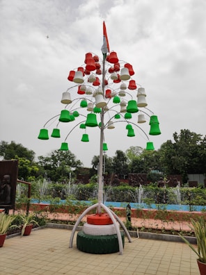 A creatively designed artificial tree made up of colorful pot-like structures in red, white, and green is displayed in an outdoor setting. It is topped with a national flag. The background features lush greenery, fountains, and sculpted panels. Potted plants are arranged at the base, while the sky appears overcast.
