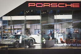 A showroom with glass walls displaying several luxury cars, including a silver sports car in the foreground and a white vehicle behind it. The interior features modern furniture like brown chairs and decorative plants, with a sleek, contemporary design. The scene is well-lit, highlighting the glossy surfaces and creating an atmosphere of sophistication and elegance. The word 'Porsche' is prominently displayed at the top.