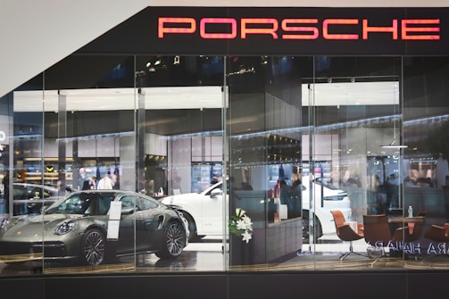 A showroom with glass walls displaying several luxury cars, including a silver sports car in the foreground and a white vehicle behind it. The interior features modern furniture like brown chairs and decorative plants, with a sleek, contemporary design. The scene is well-lit, highlighting the glossy surfaces and creating an atmosphere of sophistication and elegance. The word 'Porsche' is prominently displayed at the top.