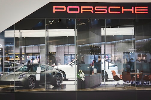 A showroom with glass walls displaying several luxury cars, including a silver sports car in the foreground and a white vehicle behind it. The interior features modern furniture like brown chairs and decorative plants, with a sleek, contemporary design. The scene is well-lit, highlighting the glossy surfaces and creating an atmosphere of sophistication and elegance. The word 'Porsche' is prominently displayed at the top.