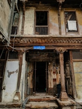 A weathered building façade featuring intricate wooden carvings and pillars. The structure looks aged, with peeling paint and visible wear. The sign reading 'Unit Trust of India' hangs above the entrance, suggesting a historical or repurposed location. There are barred windows and open doorways, with some clutter collected around the entrance.
