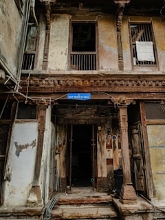 A weathered building façade featuring intricate wooden carvings and pillars. The structure looks aged, with peeling paint and visible wear. The sign reading 'Unit Trust of India' hangs above the entrance, suggesting a historical or repurposed location. There are barred windows and open doorways, with some clutter collected around the entrance.