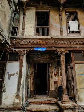 A weathered building façade featuring intricate wooden carvings and pillars. The structure looks aged, with peeling paint and visible wear. The sign reading 'Unit Trust of India' hangs above the entrance, suggesting a historical or repurposed location. There are barred windows and open doorways, with some clutter collected around the entrance.