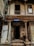A weathered building façade featuring intricate wooden carvings and pillars. The structure looks aged, with peeling paint and visible wear. The sign reading 'Unit Trust of India' hangs above the entrance, suggesting a historical or repurposed location. There are barred windows and open doorways, with some clutter collected around the entrance.