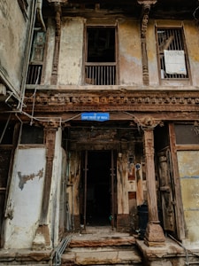 A weathered building façade featuring intricate wooden carvings and pillars. The structure looks aged, with peeling paint and visible wear. The sign reading 'Unit Trust of India' hangs above the entrance, suggesting a historical or repurposed location. There are barred windows and open doorways, with some clutter collected around the entrance.