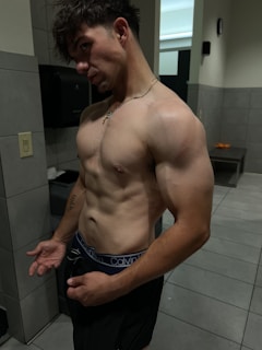 A shirtless man with muscular build standing in a gym locker room. The lighting highlights his physique, and he is wearing dark shorts with visible brand underwear.
